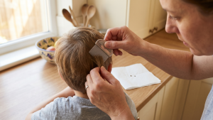 A mother in a sunny home kitchen carefully combs through her young son's hair with a fine-toothed metal nit comb during a post-treatment check, with two suspect specks placed beside her on a folded white paper towel for inspection.